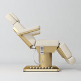 Manufakturo Aurélie beauty bed in Creamy with gold-finished base, side view with backrest raised and leg section lowered.