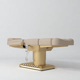 Manufakturo Aurélie beauty bed in Warm Beige with gold-finished base, flat low-angle view.
