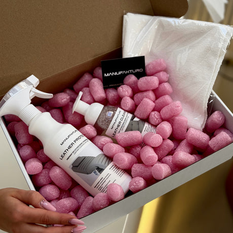 A hand holds an open cardboard box filled with pink packing peanuts and a white microfiber cloth, containing three white bottles of Manufakturo leather care products: a leather cleaner, a leather protector spray, and a leather conditioner.