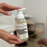 A person holds a bottle of leather cleaner with a pump dispenser. The bottle is clear, showing a beige liquid inside, and has a white pump and a white label with black text.