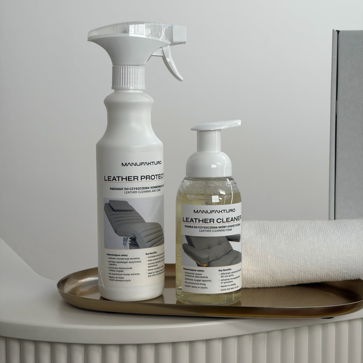 Two Manufakturo Leather Care products, a spray bottle and a pump bottle of leather cleaner, displayed on a gold tray next to a rolled white towel.