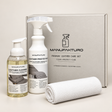 Manufakturo premium leather care set including a leather cleaner pump bottle, a leather protector spray bottle, a rolled white microfiber cloth, and a gray presentation box on a light beige background.