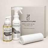 Manufakturo premium leather care set including a leather cleaner pump bottle, a leather protector spray bottle, a rolled white microfiber cloth, and a gray presentation box on a light beige background.