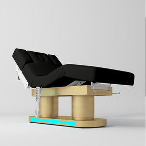 An adjustable medical or spa treatment table with a black padded bed frame, a gold support base, and a white control panel, shown on a light gray studio background.
