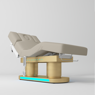 An electric treatment bed with a grey-colored padded frame and a gold-colored base, shown on a light gray studio background.