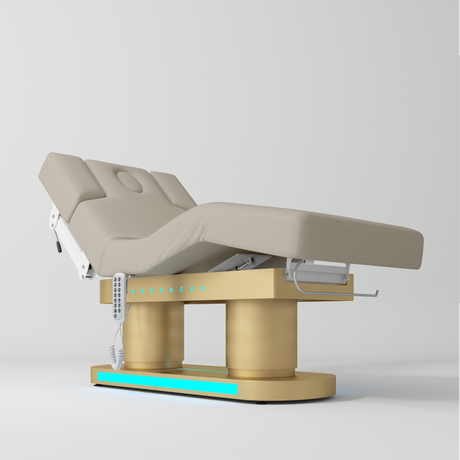 An electric treatment bed with a grey-colored padded frame and a gold-colored base, shown on a light gray studio background.