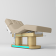 An adjustable electric treatment bed with a beige padded frame and base, featuring a control panel and white support arms, shown on a light gray studio background.