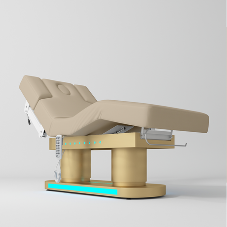 An adjustable electric treatment bed with a beige padded frame and base, featuring a control panel and white support arms, shown on a light gray studio background.