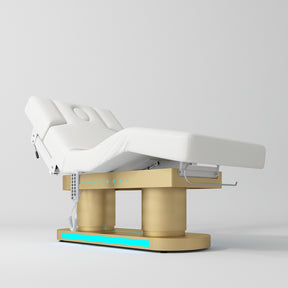 An adjustable medical examination table with a white padded surface and a gold-colored metal base, shown on a clean light gray studio background.