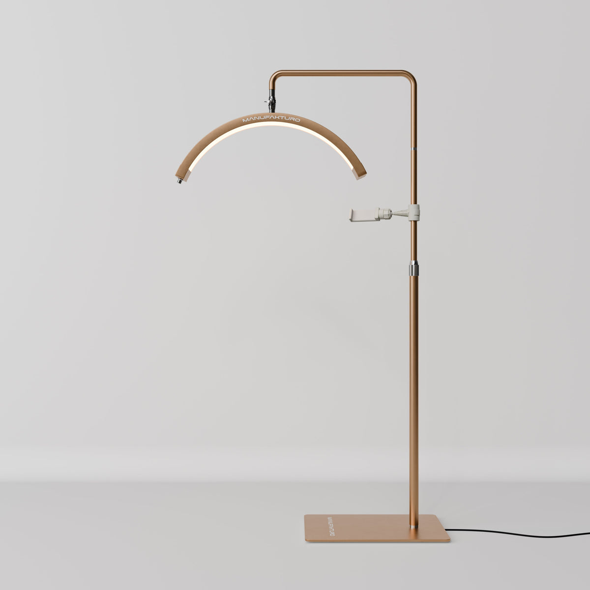 Original product photograph of the Manufakturo V3 Pro Gold treatment lamp, showing the actual gold finish, arched LED head, phone holder and adjustable stand.