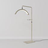 A modern floor lamp with a slim, adjustable stand and an arched light bar, finished in a warm cream or beige tone. The lamp is shown against a clean, light gray studio background with its power cord visible on the right.