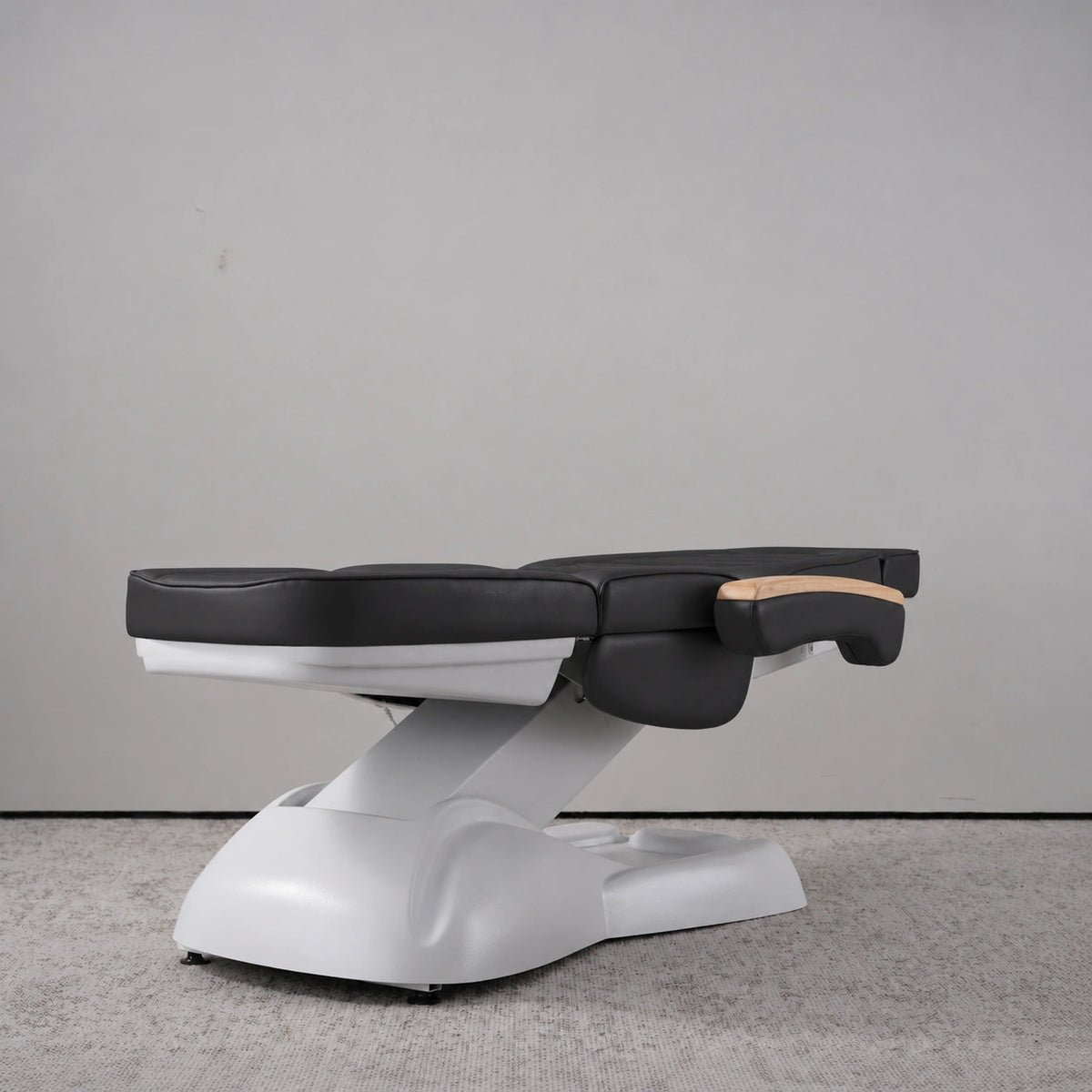 Manufakturo Grace Black electric beauty bed lying flat, with wooden armrest caps and the original white inclined lift and sculpted base.