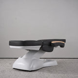 Manufakturo Grace Black electric beauty bed lying flat, with wooden armrest caps and the original white inclined lift and sculpted base.