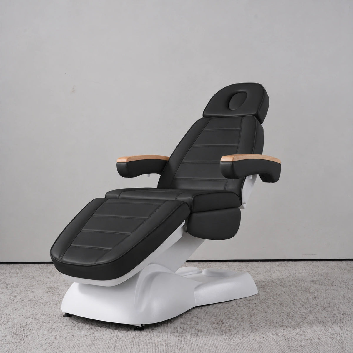 Manufakturo Grace Black electric beauty bed in a treatment position, with wooden armrest caps and a white sculpted base.