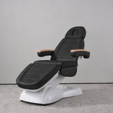 Manufakturo Grace Black electric beauty bed in a treatment position, with wooden armrest caps and a white sculpted base.