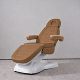 Manufakturo Grace Caramel electric beauty bed in a treatment position, with wooden armrest caps and a white sculpted base.