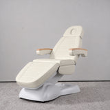 Manufakturo Grace Creamy electric beauty bed in a treatment position, with wooden armrest caps and a white sculpted base.