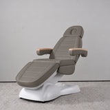 Manufakturo Grace Deep Ash electric beauty bed in a treatment position, with wooden armrest caps and a white sculpted base.