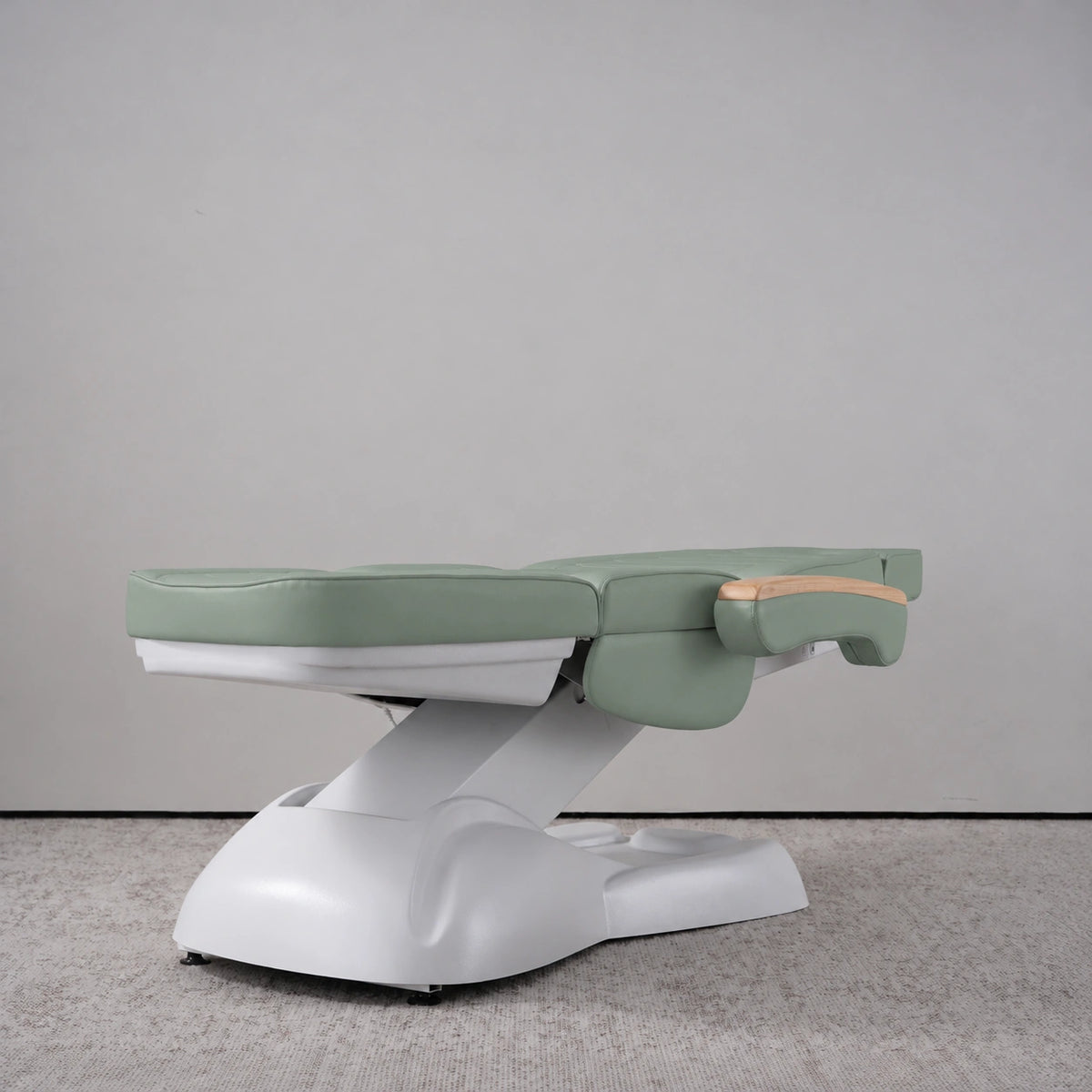 Manufakturo Grace Olive electric beauty bed lying flat, with wooden armrest caps and the original white inclined lift and sculpted base.