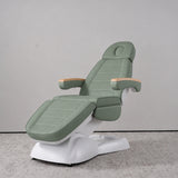 Manufakturo Grace Olive electric beauty bed in a treatment position, with wooden armrest caps and a white sculpted base.