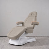 Manufakturo Grace Stone Taupe electric beauty bed in a treatment position, with wooden armrest caps and a white sculpted base.