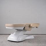 Manufakturo Grace Warm Beige electric beauty bed lying flat, with wooden armrest caps and the original white inclined lift and sculpted base.