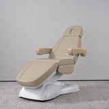 Manufakturo Grace Warm Beige electric beauty bed in a treatment position, with wooden armrest caps and a white sculpted base.