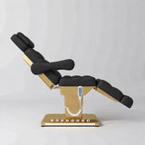 Manufakturo Orielle B Black electric beauty bed shown from the side in a treatment position, with gold base and hand-held controller.