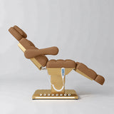 Manufakturo Orielle B Caramel electric beauty bed shown from the side in a treatment position, with gold base and hand-held controller.