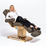 Manufakturo Orielle B Cocoa Mocha beauty bed in use, showing upholstered armrests, extendable footrest, hand and foot controls and LED base lighting.
