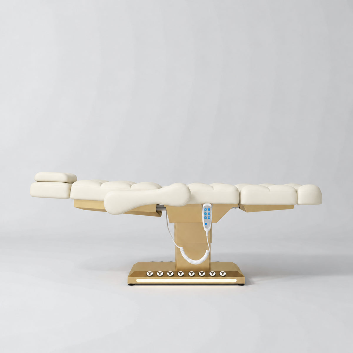 Manufakturo Orielle B Creamy electric beauty bed in a flat position viewed from the side, with gold base and hand-held controller.