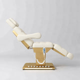 Manufakturo Orielle B Creamy electric beauty bed shown from the side in a treatment position, with gold base and hand-held controller.