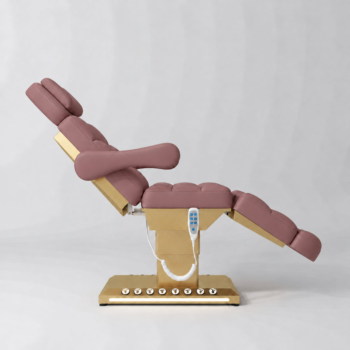 Manufakturo Orielle B Dusty Rose electric beauty bed shown from the side in a treatment position, with gold base and hand-held controller.