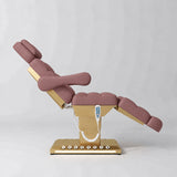 Manufakturo Orielle B Dusty Rose electric beauty bed shown from the side in a treatment position, with gold base and hand-held controller.