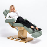 Manufakturo Orielle B Olive beauty bed in use, showing upholstered armrests, extendable footrest, hand and foot controls and LED base lighting.