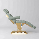 Manufakturo Orielle B Olive electric beauty bed shown from the side in a treatment position, with gold base and hand-held controller.