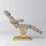 Manufakturo Orielle B Pearl Greige electric beauty bed shown from the side in a treatment position, with gold base and hand-held controller.