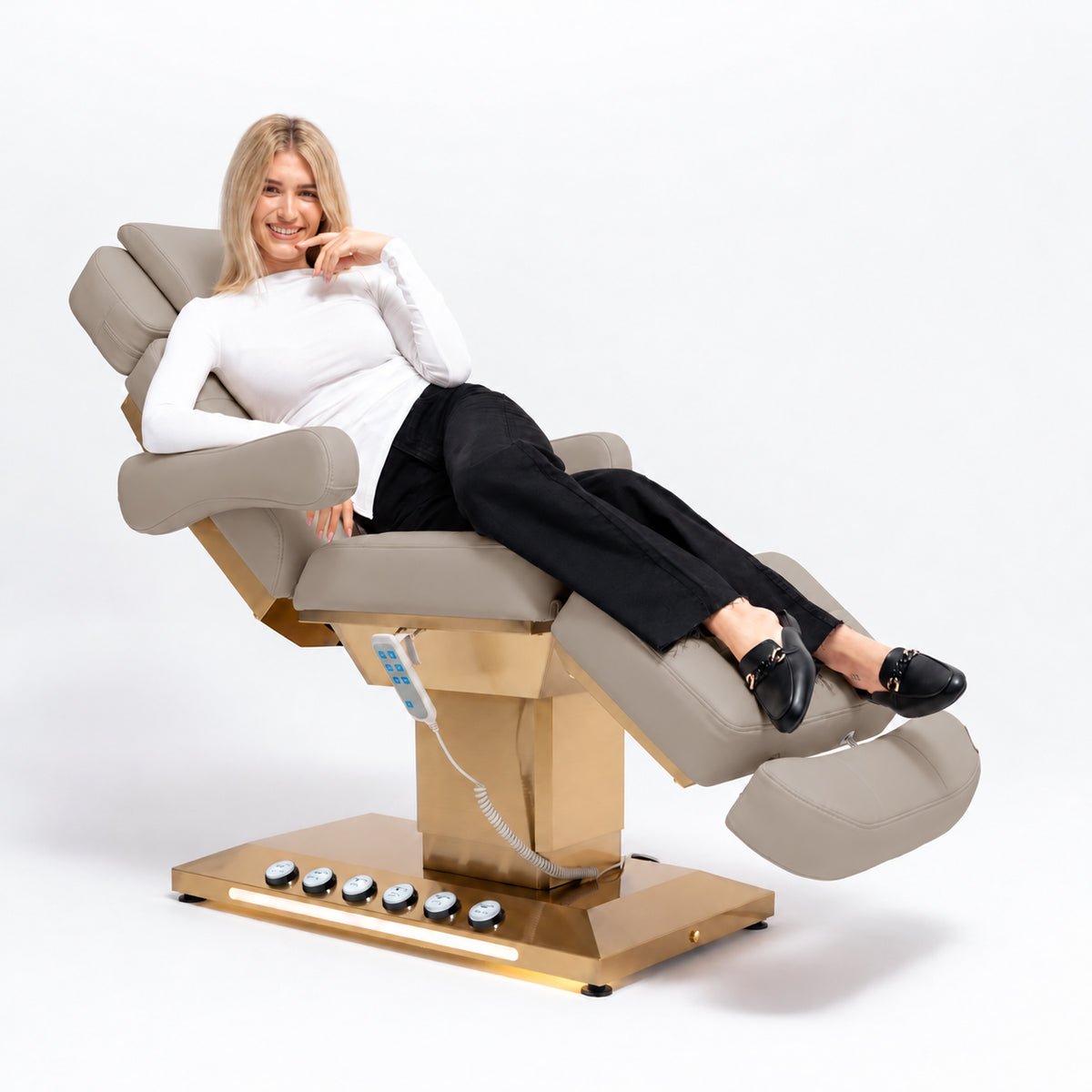 Manufakturo Orielle B Stone Taupe beauty bed in use, showing upholstered armrests, extendable footrest, hand and foot controls and LED base lighting.