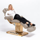 Manufakturo Orielle B Stone Taupe beauty bed in use, showing upholstered armrests, extendable footrest, hand and foot controls and LED base lighting.