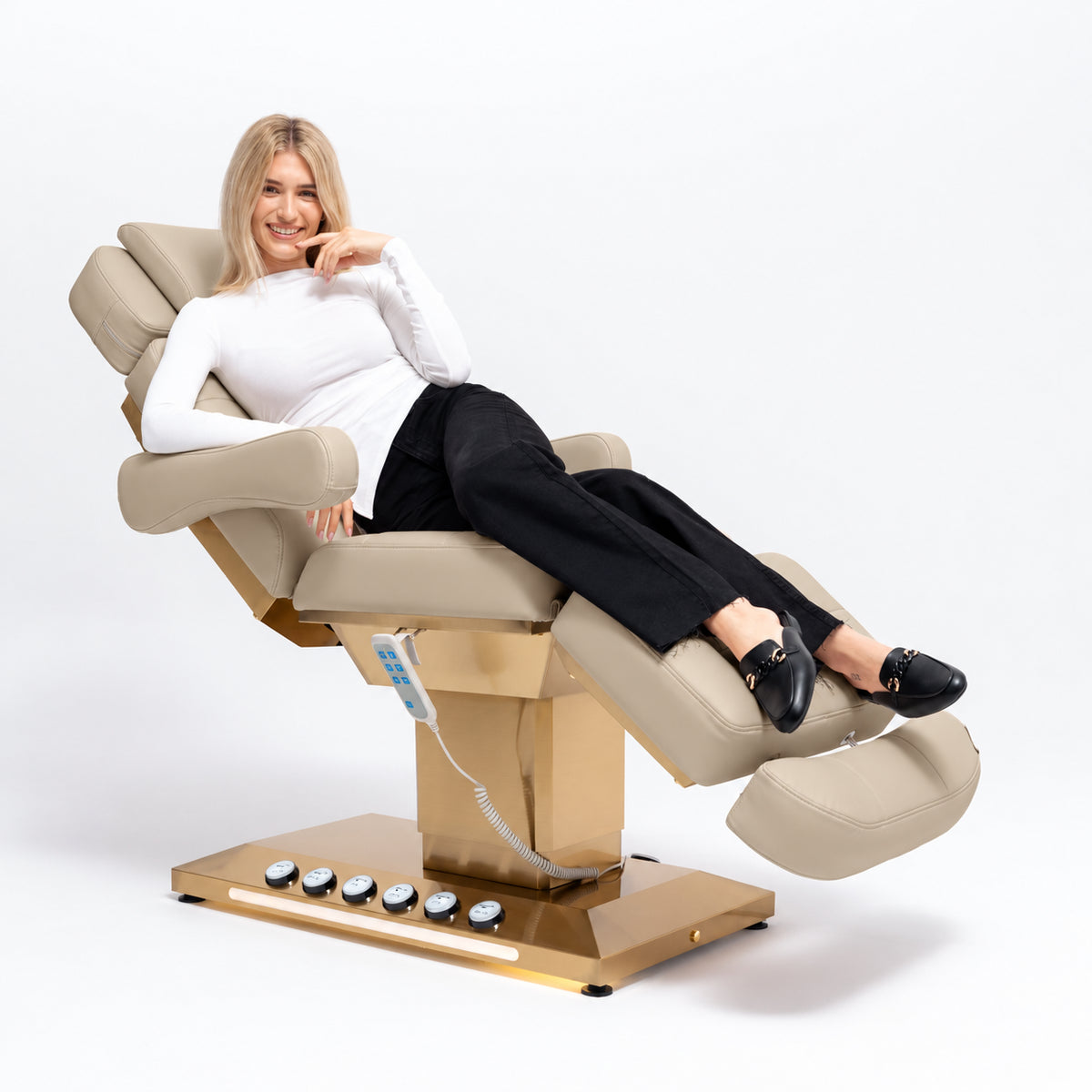 Manufakturo Orielle B Warm Beige beauty bed in use, showing upholstered armrests, extendable footrest, hand and foot controls and LED base lighting.