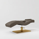 Manufakturo Prestige Cocoa Mocha hydraulic eyelash bed, three-quarter side view with gold pedestal base and contoured upholstery.