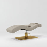 Manufakturo Prestige Stone Taupe hydraulic eyelash bed, three-quarter side view with gold pedestal base and contoured upholstery.