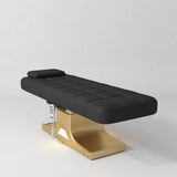 Manufakturo Saphir A Black electric beauty bed with gold base, flat lying position shown in a three-quarter view, without armrests.