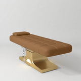 Manufakturo Saphir A Caramel electric beauty bed with gold base, flat lying position shown in a three-quarter view, without armrests.