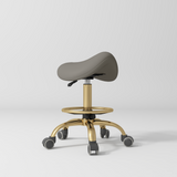Manufakturo Selle Ergonomic Beauty Saddle Stool Deep Ash - Gold Base - displayed in a professional treatment-room setting