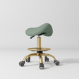 Manufakturo Selle Ergonomic Beauty Saddle Stool Olive - Gold Base - displayed in a professional treatment-room setting