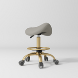 Manufakturo Selle Ergonomic Beauty Saddle Stool Pearl Greige - Gold Base - displayed in a professional treatment-room setting