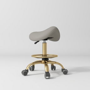 Manufakturo Selle Ergonomic Beauty Saddle Stool Pearl Greige - Gold Base - displayed in a professional treatment-room setting