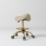 Manufakturo Selle Ergonomic Beauty Saddle Stool Warm Beige - Gold Base - displayed in a professional treatment-room setting