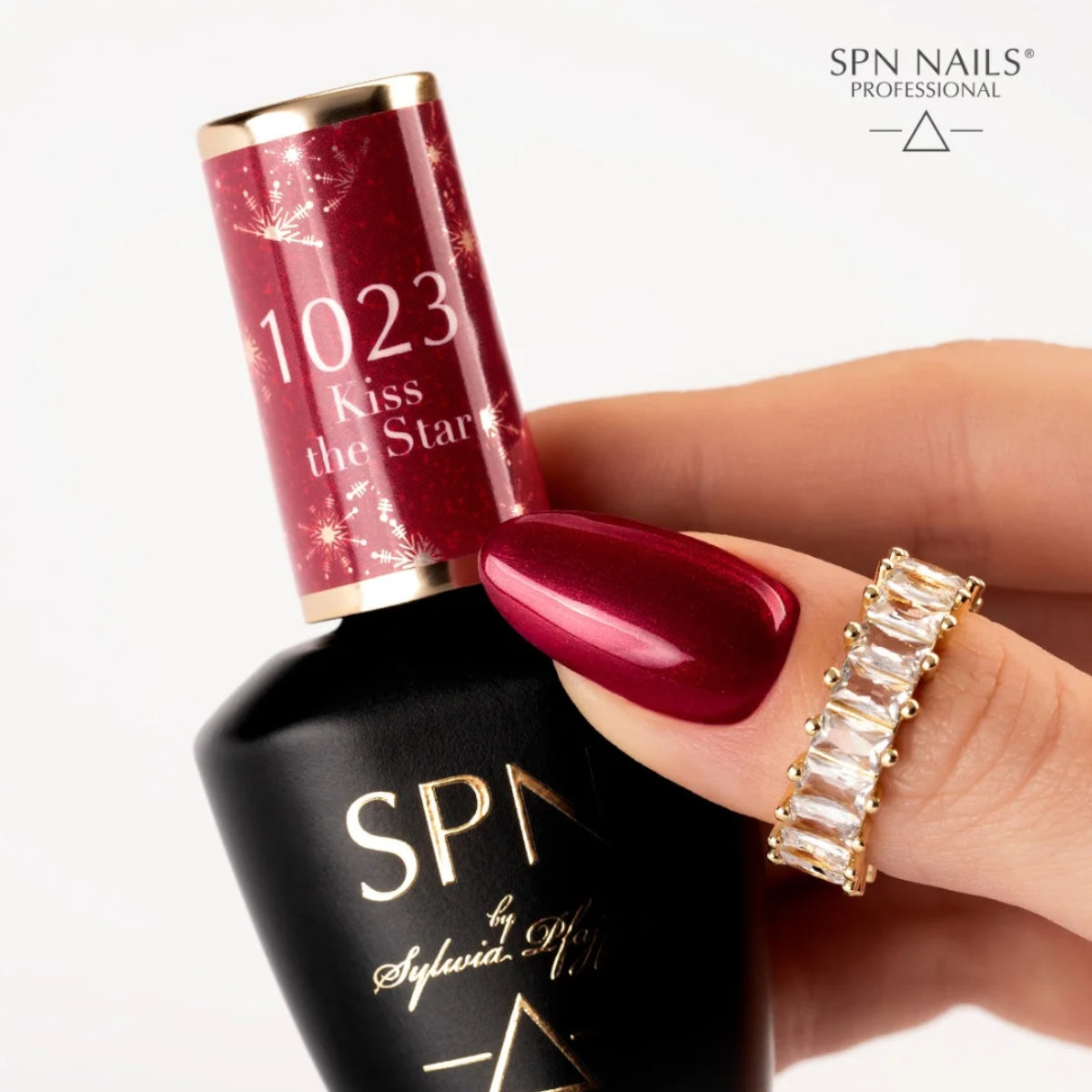 SPN Nails UV/LED Gel Polish 1023 Kiss the Star bottle held by a hand with red glitter nails, paired with a gold ring.